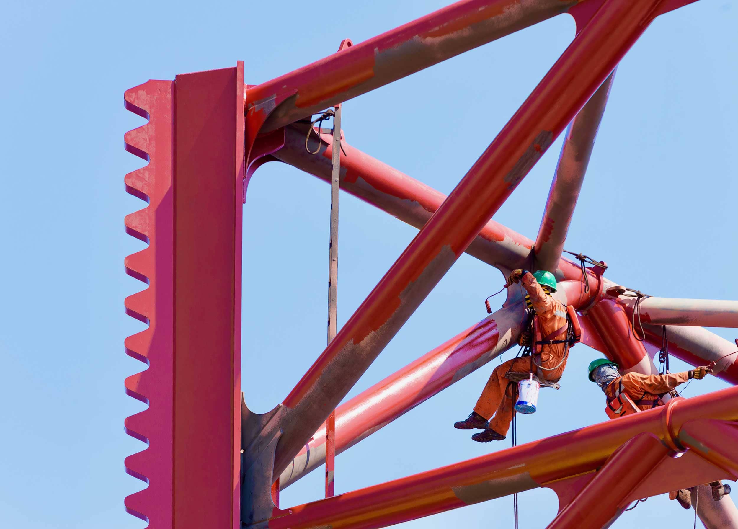 Workers in safety gear carefully paint and maintain the legs of a newbuild WTIV under a clear blue sky.